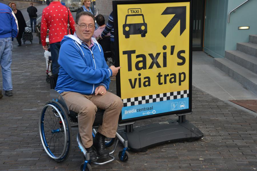 Man in een rolstoel wijst naar bord waarop staat, Taxi's via de trap omhoog