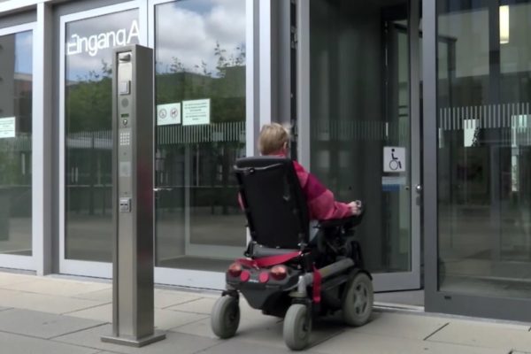 Een foto waarin een man in een elektrische rolstoel een goed toegankelijke ingang van een gebouw binnenrijdt.